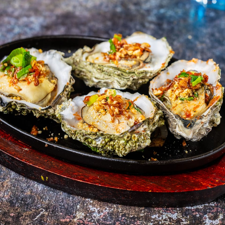 Four Vietnamese grilled oysters topped with crispy shallots and green onions on a black sizzling plate.