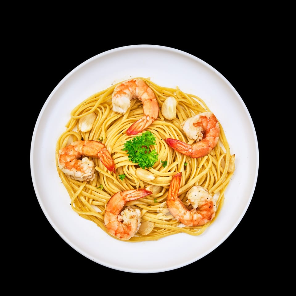 A white plate of spaghetti with shrimp, garlic cloves, and a sprig of parsley.