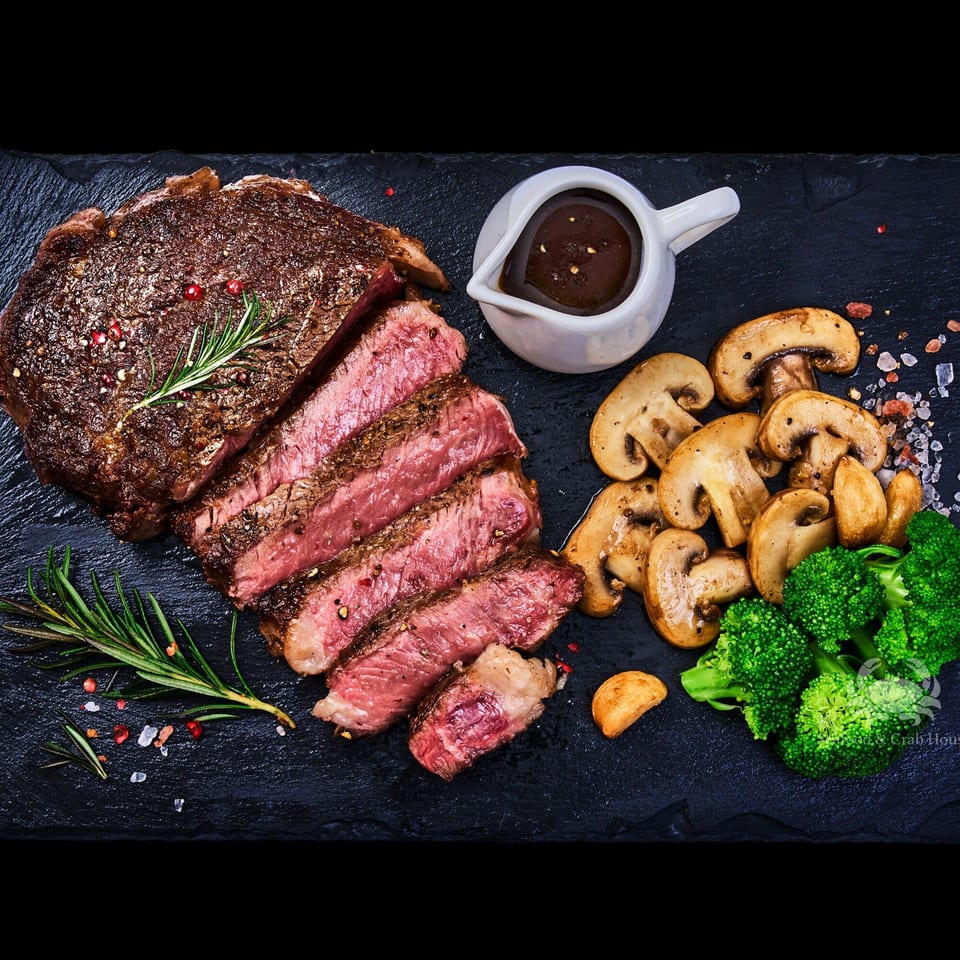 Sliced steak with rosemary, mushrooms, broccoli, and a small pitcher of sauce on a dark platter.